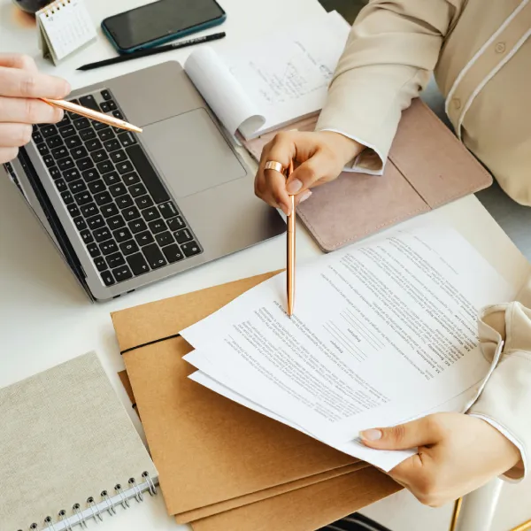 Two people pointing at a document on a desk with a laptop and folders